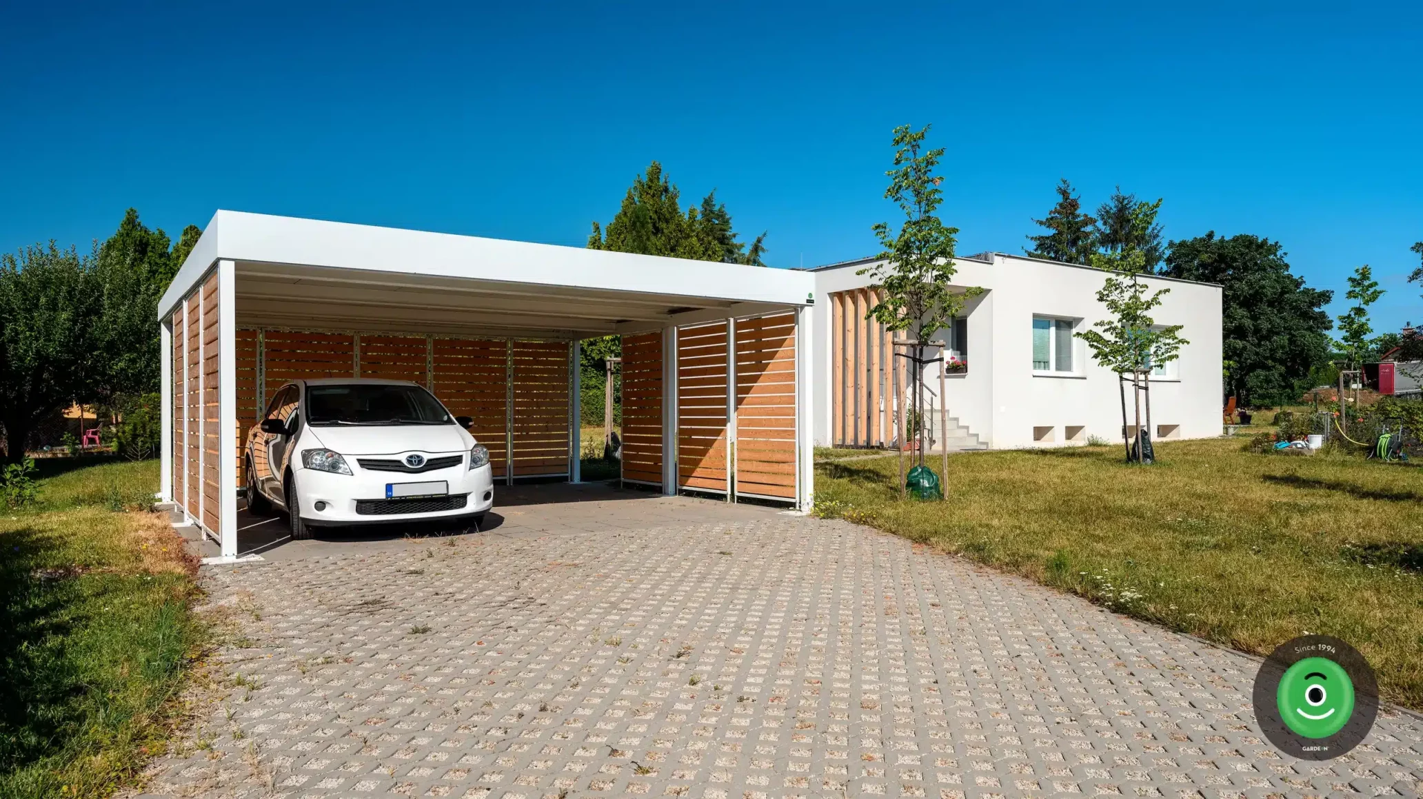Přístřešek GARDEON s bílou střechou u bílého rodinného domu.