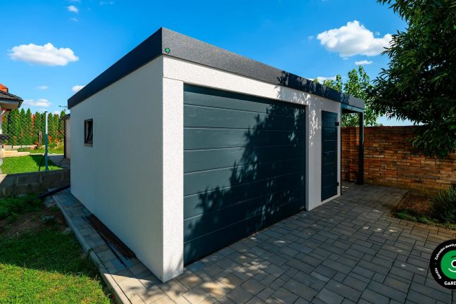 Bílá montovaná garáž s tmavě šedou bránou a dveřmi, elegantní vzhled na dvoře rodinného domu.